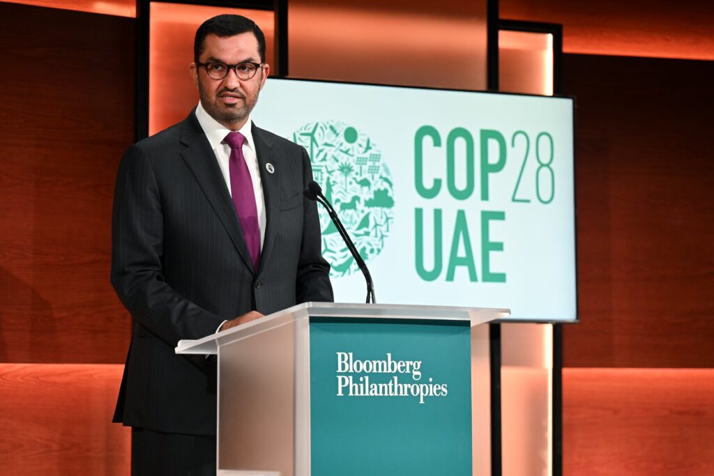 Sultan Al Jaber stands at a podium, speaking, with a COP28 sign behind him on stage
