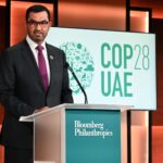 COP in crisis Sultan Al Jaber stands at a podium, speaking, with a COP28 sign behind him on stage
