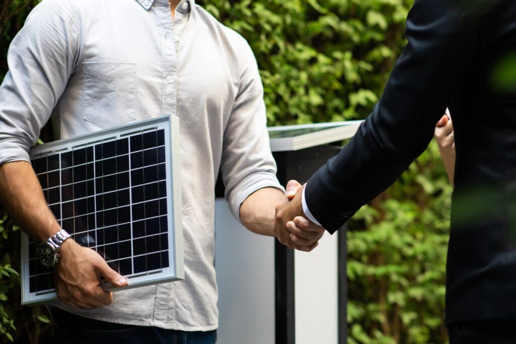 Two men in semi-professional attire shake hands, while one is holding a small sample of solar panel technology