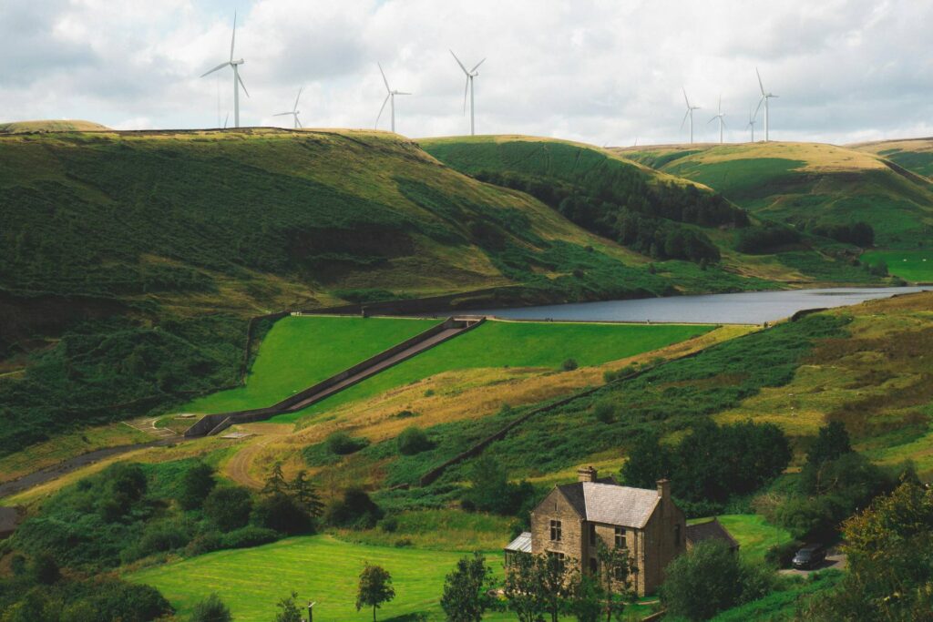 View of the Greenbooth Reservoir, Greater Manchester, England. A stone house sits by the reservoir with wind turbines on the ridge in the distance.