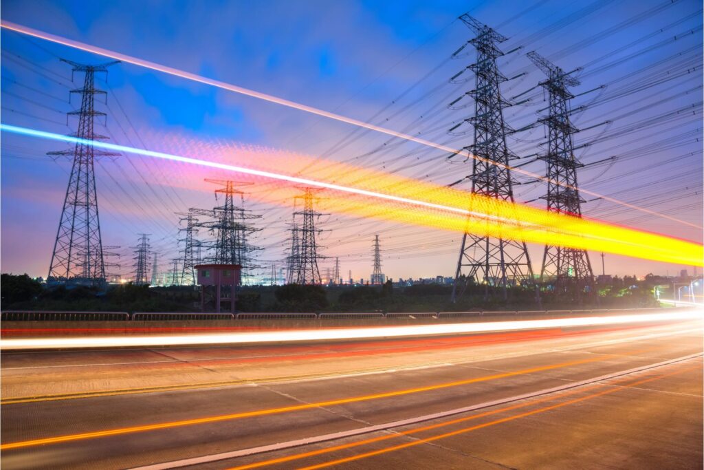 Car lights blur in front of power lines as the sun sets in the distance.