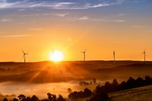 The sun sets on the horizon, outlining wind turbines on the crest of a hill