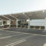 Government considers mandating solar PV in car parks A white car sits under a solar carport on a sunny day.