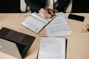 Professional person writing in a journal on a desk. A laptop and other shets of paper are also on the desk.